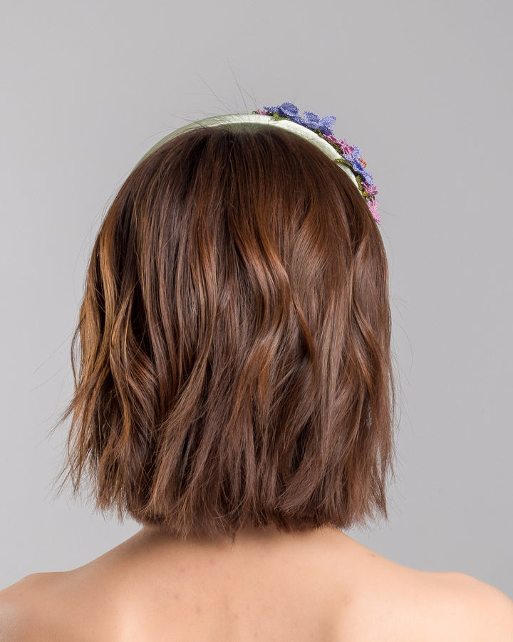 The model is showcasing the back side close up of the sage green satin padded headband embellished with handcrafted Turkish needle embroidery flowers in lilac and pink tones, featuring detailed petals and green stitched leaves clustered along the top of the band.  Designed and made by Merve Bayindir Millinery.
