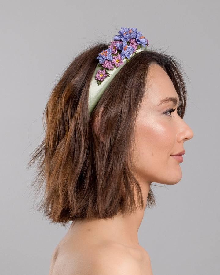 The model is showcasing the right side close up of the sage green satin padded headband embellished with handcrafted Turkish needle embroidery flowers in lilac and pink tones, featuring detailed petals and green stitched leaves clustered along the top of the band.  Designed and made by Merve Bayindir Millinery.
