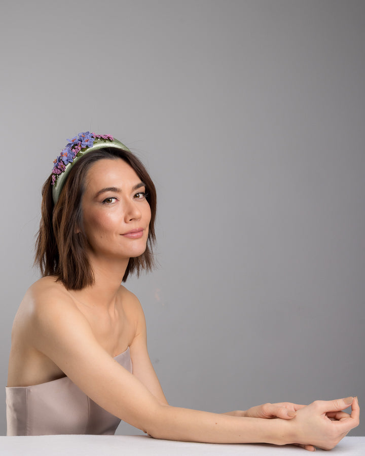 The female model is posing, wearing Lillie,  the sage green satin padded headband embellished with handcrafted Turkish needle embroidery flowers in lilac and pink tones, featuring detailed petals and green stitched leaves clustered along the top of the band.  Designed and made by Merve Bayindir Millinery.
