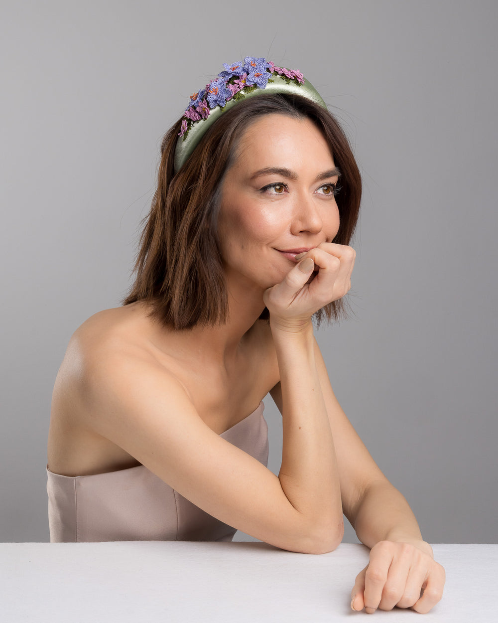 The female model is posing, wearing Lillie,  the sage green satin padded headband embellished with handcrafted Turkish needle embroidery flowers in lilac and pink tones, featuring detailed petals and green stitched leaves clustered along the top of the band.  Designed and made by Merve Bayindir Millinery.