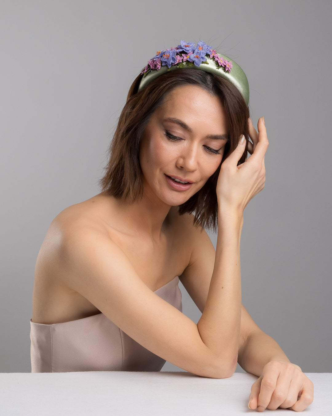 The female model is posing, wearing Lillie,  the sage green satin padded headband embellished with handcrafted Turkish needle embroidery flowers in lilac and pink tones, featuring detailed petals and green stitched leaves clustered along the top of the band.  Designed and made by Merve Bayindir Millinery.