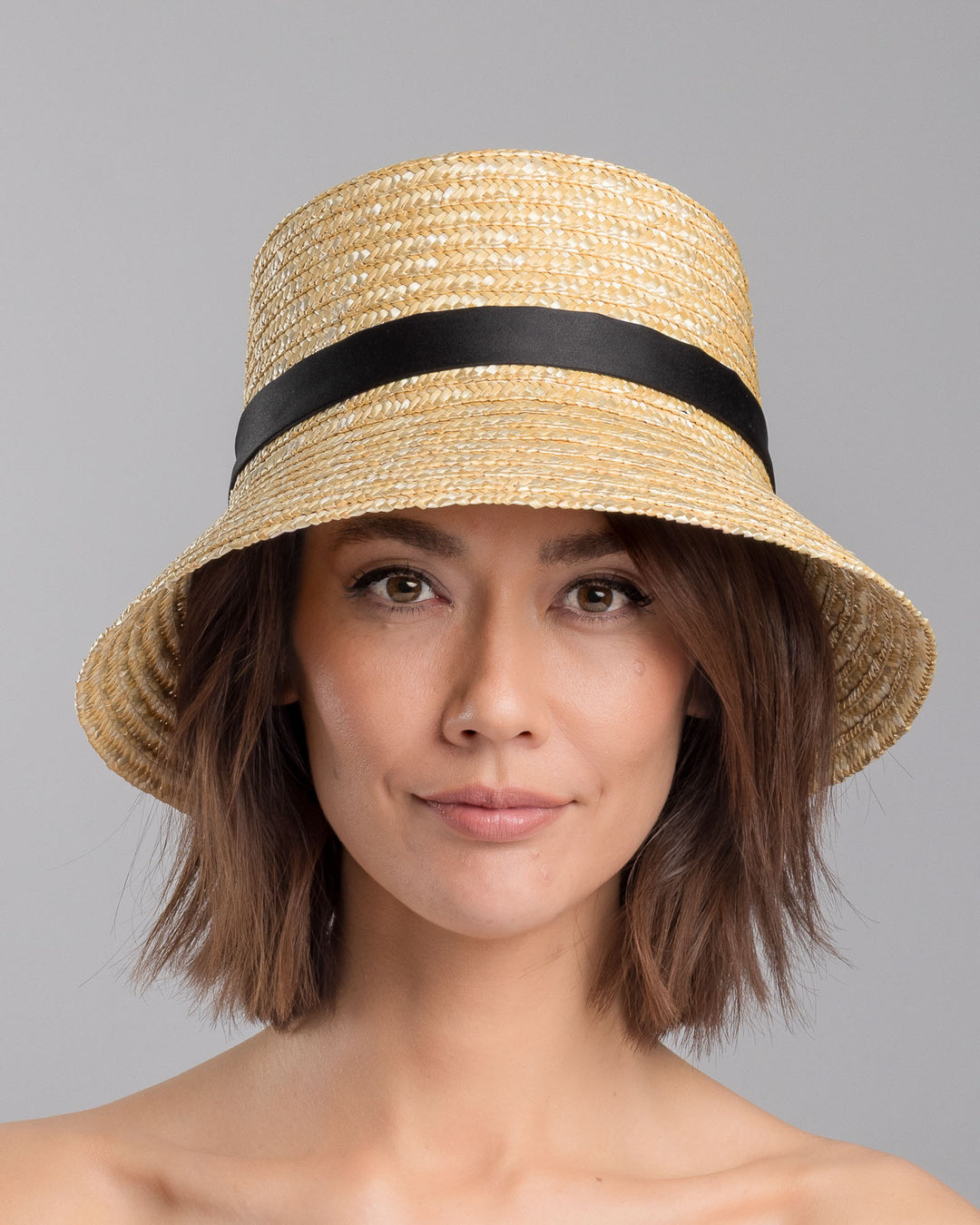 The model is showcasing the front side close up of the natural woven straw bucket hat with a softly sloping brim, finished with a black ribbon band and long trailing ties at the side. Designed and Made by Merve Bayindir millinery.