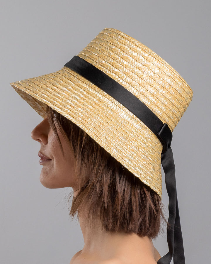 The model is showcasing the left  side close up of the natural woven straw bucket hat with a softly sloping brim, finished with a black ribbon band and long trailing ties at the side. Designed and Made by Merve Bayindir millinery.