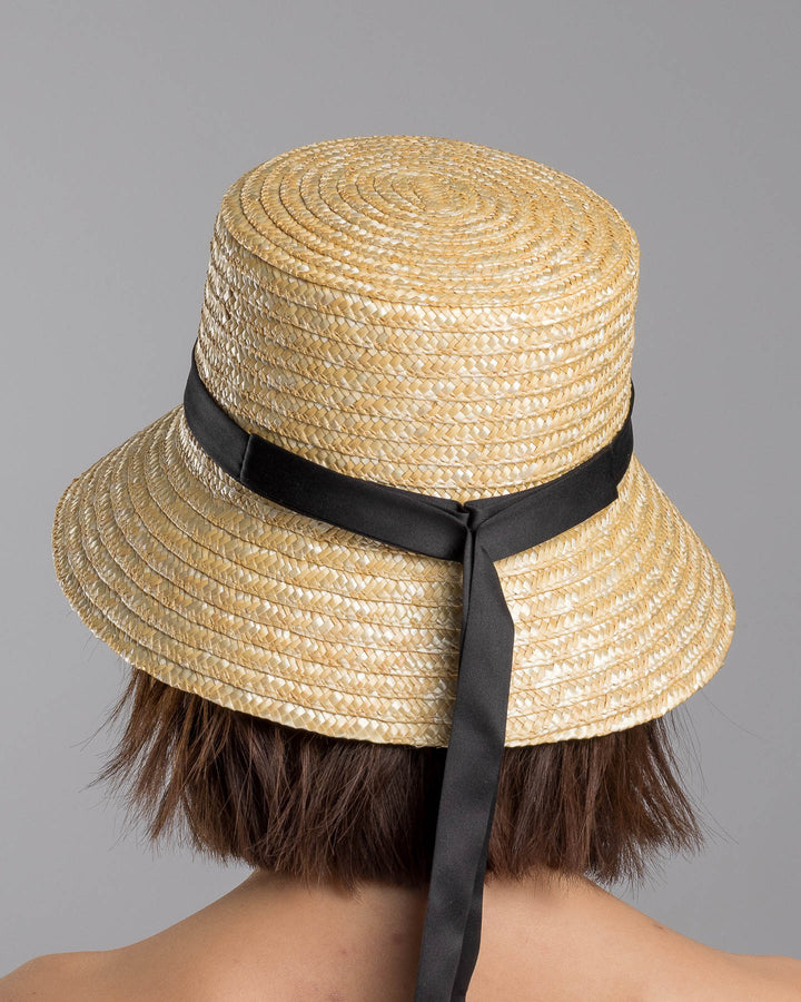 The model is showcasing the back side close up of the natural woven straw bucket hat with a softly sloping brim, finished with a black ribbon band and long trailing ties at the side. Designed and Made by Merve Bayindir millinery.