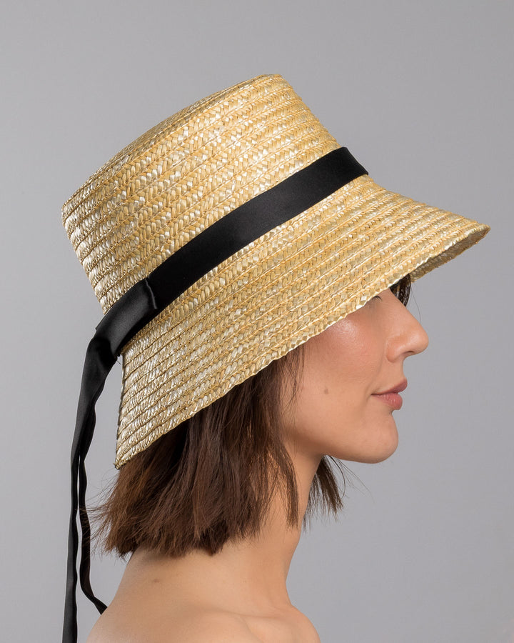 The model is showcasing the right side close up of the natural woven straw bucket hat with a softly sloping brim, finished with a black ribbon band and long trailing ties at the side. Designed and Made by Merve Bayindir millinery.