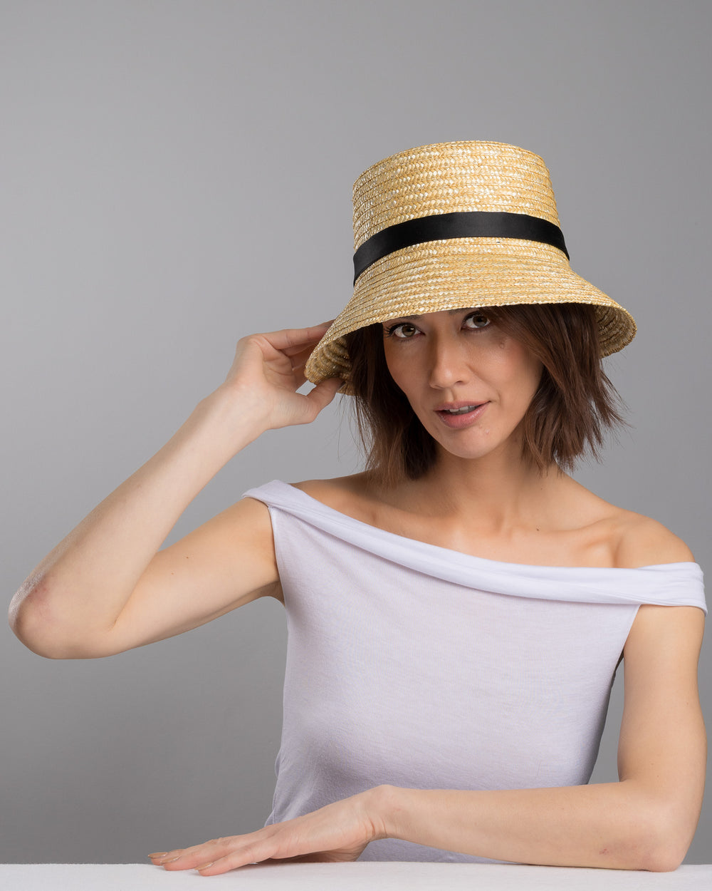 The female model is posing, wearing Lunor, the natural woven straw bucket hat with a softly sloping brim, finished with a black ribbon band and long trailing ties at the side. Designed and Made by Merve Bayindir millinery.