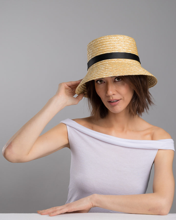The female model is posing, wearing Lunor, the natural woven straw bucket hat with a softly sloping brim, finished with a black ribbon band and long trailing ties at the side. Designed and Made by Merve Bayindir millinery.