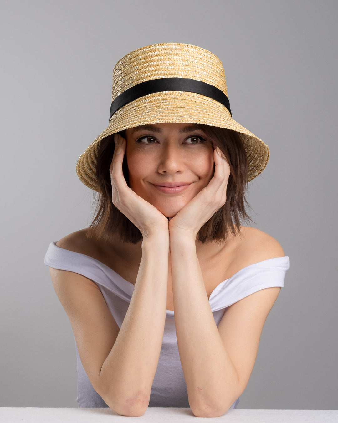 The female model is posing, wearing Lunor, the natural woven straw bucket hat with a softly sloping brim, finished with a black ribbon band and long trailing ties at the side. Designed and Made by Merve Bayindir millinery.