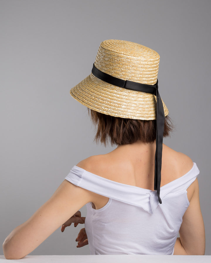 The female model is posing, wearing Lunor, the natural woven straw bucket hat with a softly sloping brim, finished with a black ribbon band and long trailing ties at the side. Designed and Made by Merve Bayindir millinery.