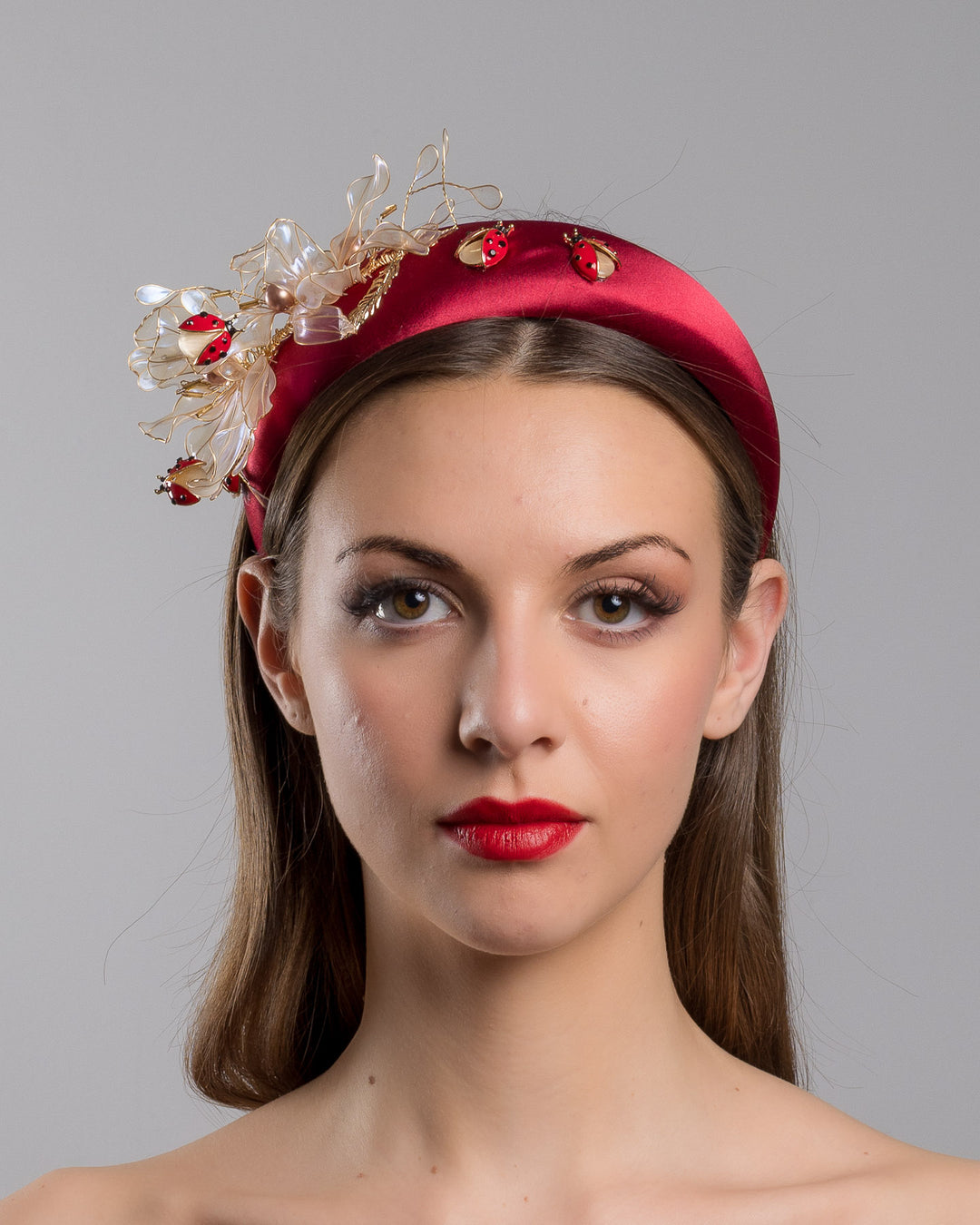 Model is showcasing the front side close up of red silk sateen headband decorated with wired resin flowers and lady bug beads. Made by Merve Bayindir Millinery.