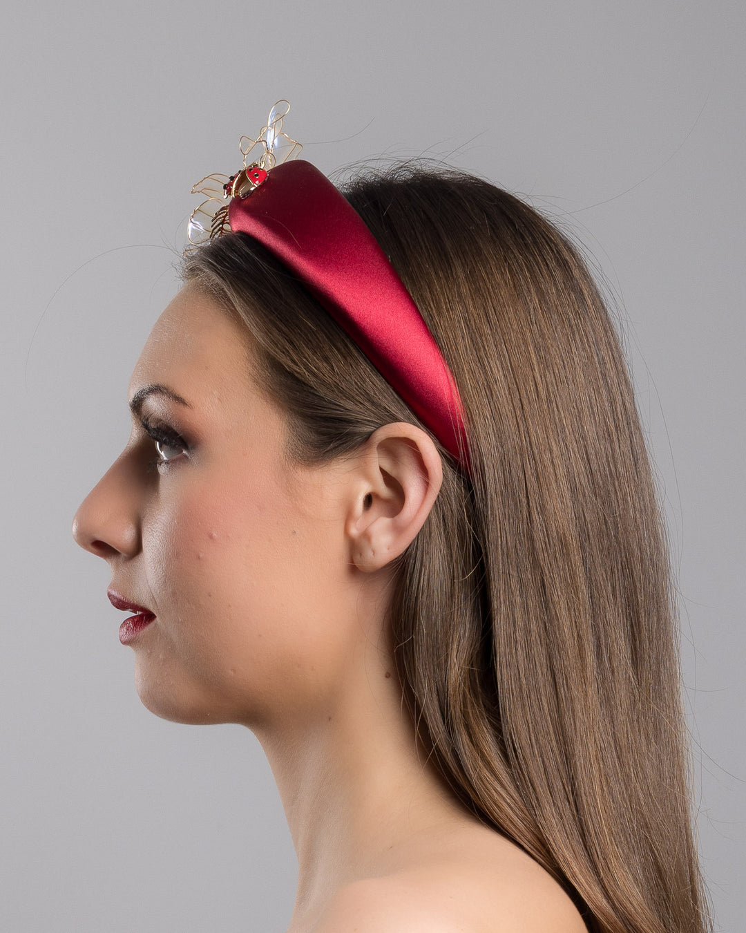 Model is showcasing the left side close up of red silk sateen headband decorated with wired resin flowers and lady bug beads. Made by Merve Bayindir Millinery.