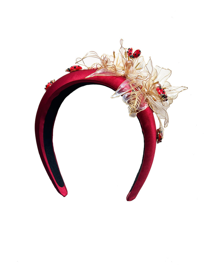 Still life picture of red silk sateen headband decorated with wired resin flowers and lady bug beads. Made by Merve Bayindir Millinery.