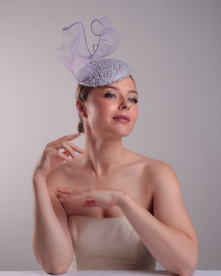 Model is showcasing the front right side of the lilac button shape fascinator covered with sequined lilac to blue mixes sequins. The top of the fascinator is adorned with a symetric flying like shape crinoline bow.