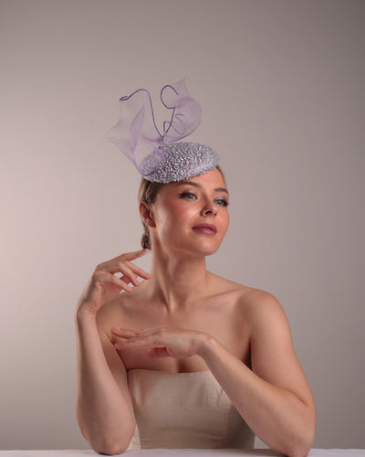 Model is showcasing the front right side of the lilac button shape fascinator covered with sequined lilac to blue mixes sequins. The top of the fascinator is adorned with a symetric flying like shape crinoline bow.