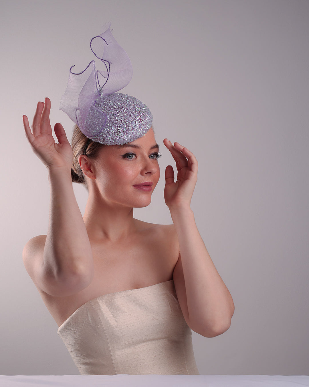Model is showcasing the front right angle of the lilac button shape fascinator covered with sequined lilac to blue mixes sequins. The top of the fascinator is adorned with a symetric flying like shape crinoline bow.