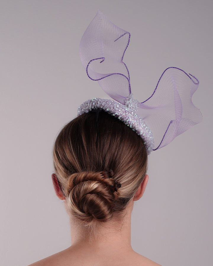 Model is showcasing the back of the lilac button shape fascinator covered with sequined lilac to blue mixes sequins. The top of the fascinator is adorned with a symetric flying like shape crinoline bow.