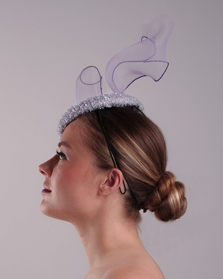 Model is showcasing the left side of the lilac button shape fascinator covered with sequined lilac to blue mixes sequins. The top of the fascinator is adorned with a symetric flying like shape crinoline bow.
