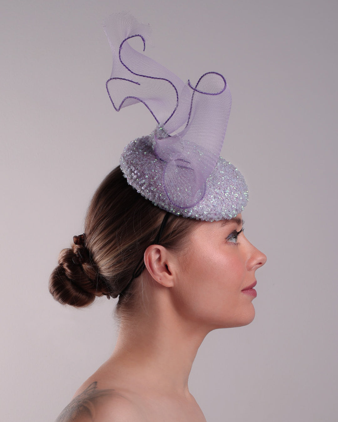 Model is showcasing the right side of the lilac button shape fascinator covered with sequined lilac to blue mixes sequins. The top of the fascinator is adorned with a symetric flying like shape crinoline bow.