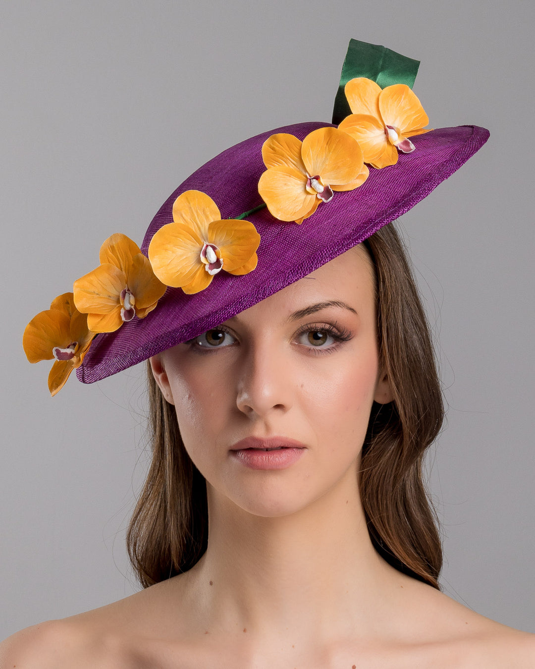 The model is showcasing the front side close up of the deep purple, round sinamay saucer hat decorated with orange orchids and a green swirl leaf. Designed and made by Merve Bayindir Millinery