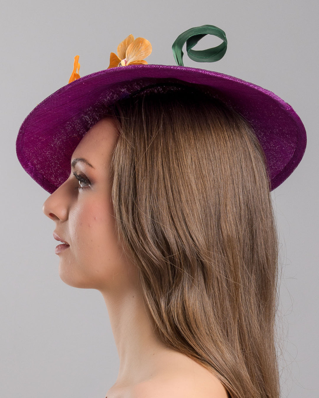 The model is showcasing the left side close up of the deep purple, round sinamay saucer hat decorated with orange orchids and a green swirl leaf. Designed and made by Merve Bayindir Millinery