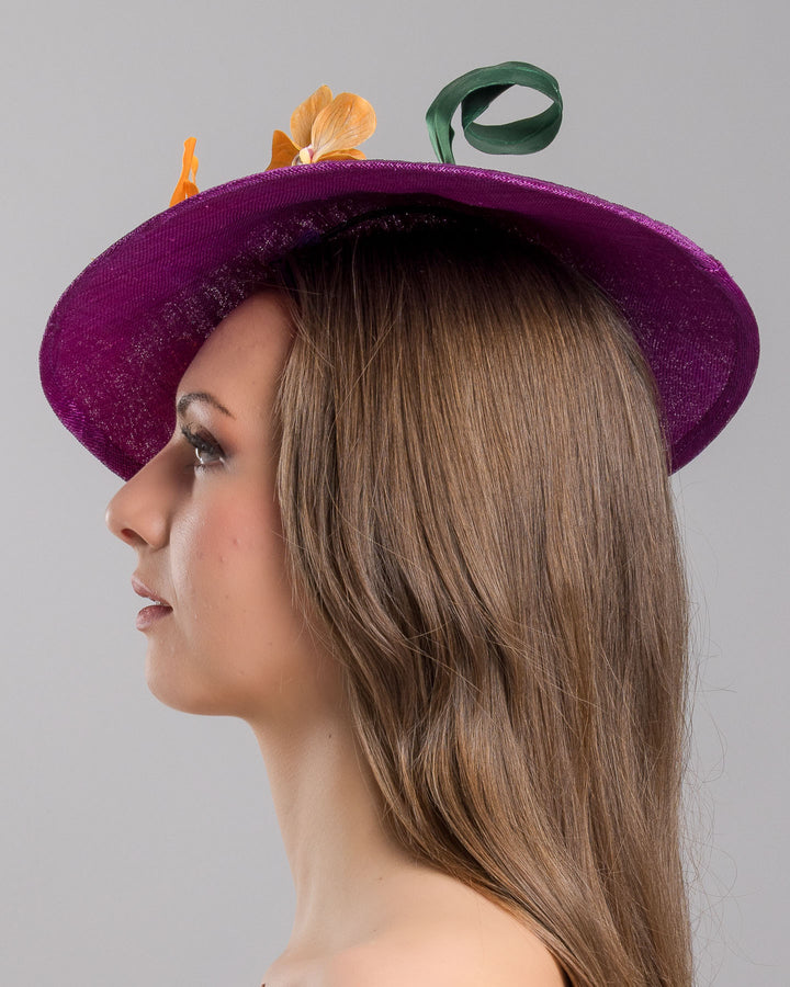 The model is showcasing the left side close up of the deep purple, round sinamay saucer hat decorated with orange orchids and a green swirl leaf. Designed and made by Merve Bayindir Millinery
