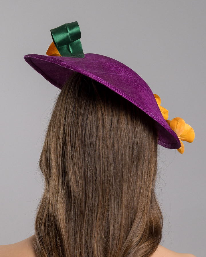 The model is showcasing the back side close up of the deep purple, round sinamay saucer hat decorated with orange orchids and a green swirl leaf. Designed and made by Merve Bayindir Millinery