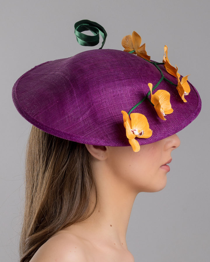 The model is showcasing the right side close up of the deep purple, round sinamay saucer hat decorated with orange orchids and a green swirl leaf. Designed and made by Merve Bayindir Millinery.
