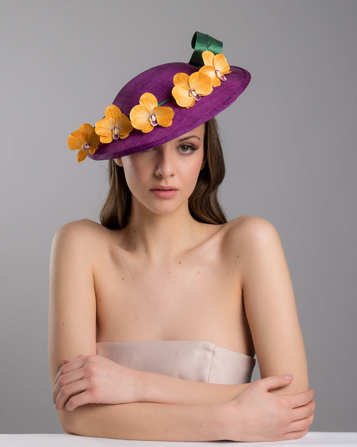 The female model is posing, wearing Lexington, the deep purple, round sinamay saucer hat decorated with orange orchids and a green swirl leaf. Designed and made by Merve Bayindir Millinery