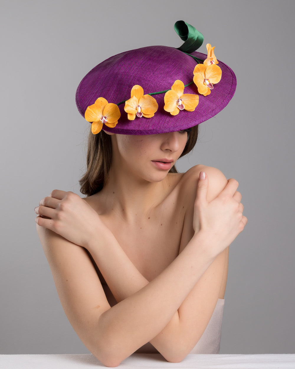 The female model is posing, wearing Lexington, the deep purple, round sinamay saucer hat decorated with orange orchids and a green swirl leaf. Designed and made by Merve Bayindir Millinery