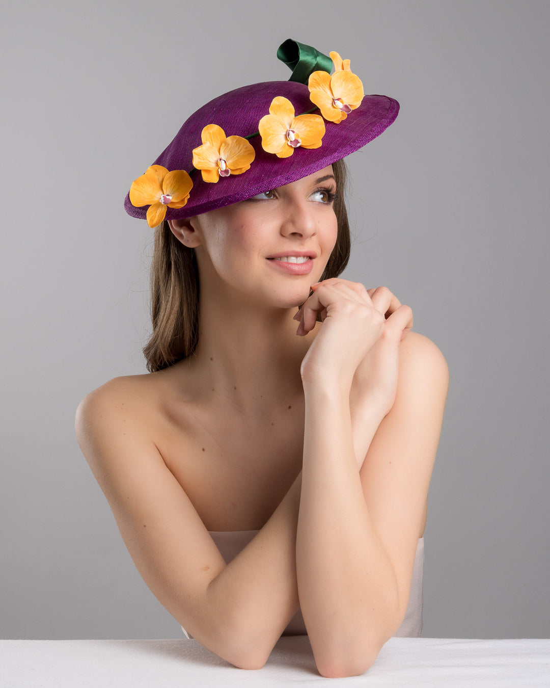The female model is posing, wearing Lexington, the deep purple, round sinamay saucer hat decorated with orange orchids and a green swirl leaf. Designed and made by Merve Bayindir Millinery