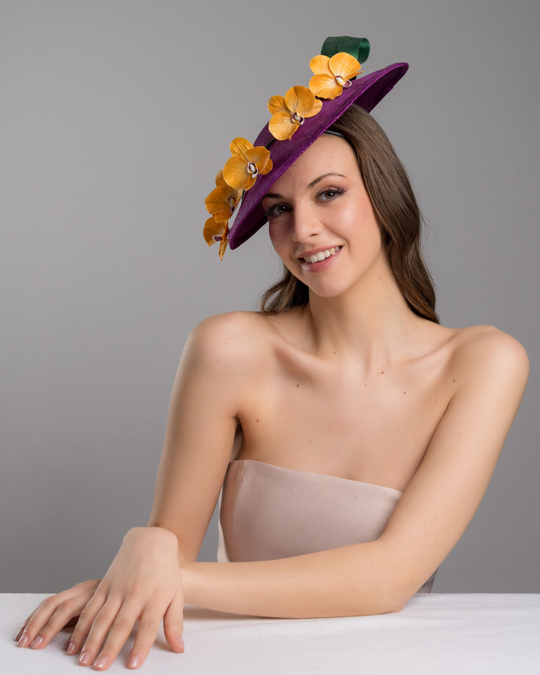 The female model is posing, wearing Lexington, the deep purple, round sinamay saucer hat decorated with orange orchids and a green swirl leaf. Designed and made by Merve Bayindir Millinery