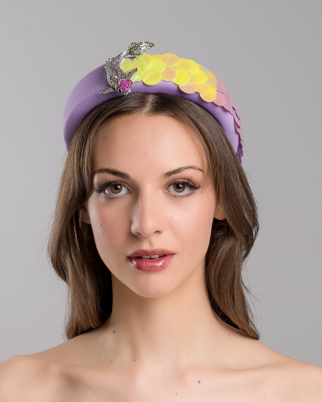 Model is showcasing the front side close up of lilac headband embroidered with green and lilac sequins on one side and embellished with a metal koi fish with crystals. Made by Merve Bayindir Millinery.