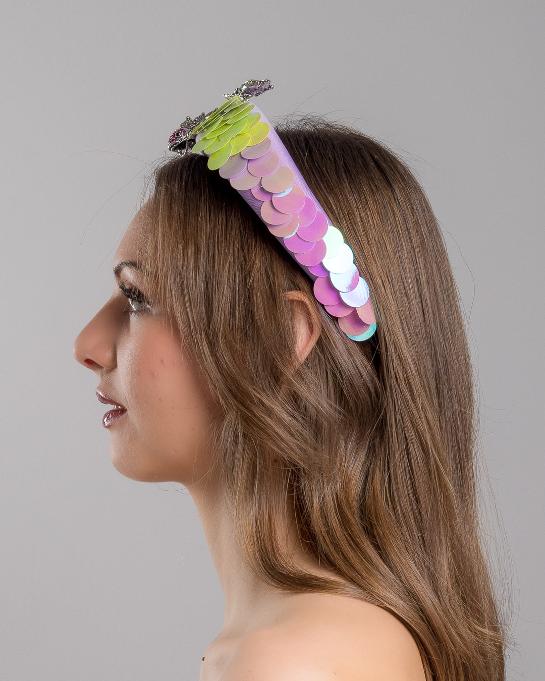 Model is showcasing the left side close up of lilac headband embroidered with green and lilac sequins on one side and embellished with a metal koi fish with crystals. Made by Merve Bayindir Millinery.