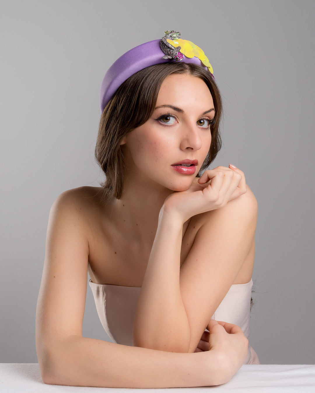 Female model is posing wearing lilac headband embroidered with green and lilac sequins on one side and embellished with a metal koi fish with crystals. Made by Merve Bayindir Millinery.
