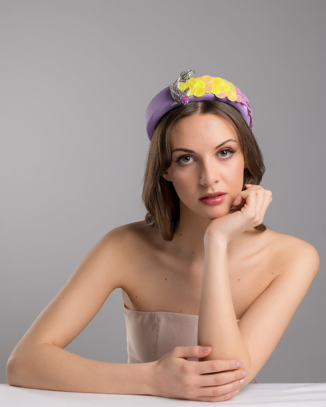 Female model is posing wearing lilac headband embroidered with green and lilac sequins on one side and embellished with a metal koi fish with crystals. Made by Merve Bayindir Millinery.