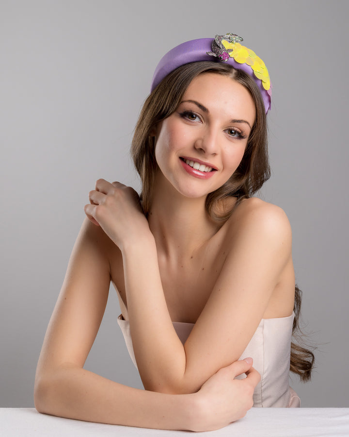 Female model is posing wearing lilac headband embroidered with green and lilac sequins on one side and embellished with a metal koi fish with crystals. Made by Merve Bayindir Millinery.