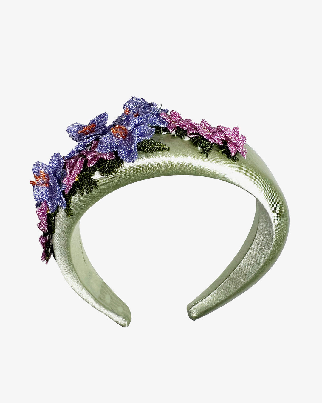 Sage green satin padded headband embellished with handcrafted Turkish needle embroidery flowers in lilac and pink tones, featuring detailed petals and green stitched leaves clustered along the top of the band.  Designed and made by Merve Bayindir Millinery.
