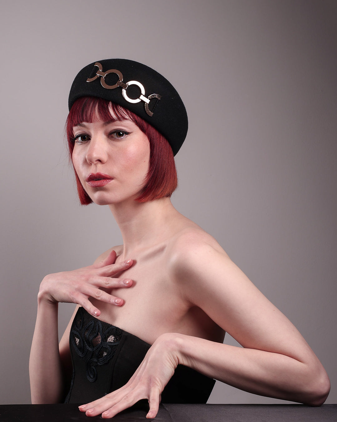 The model showcases the left front of the Liora black wool felt beret, which features a geometric gold-tone chain embellishment that arches across the crown. Designed by Merve Bayindir.