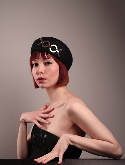 The model showcases the left front of the Liora black wool felt beret, which features a geometric gold-tone chain embellishment that arches across the crown. Designed by Merve Bayindir.