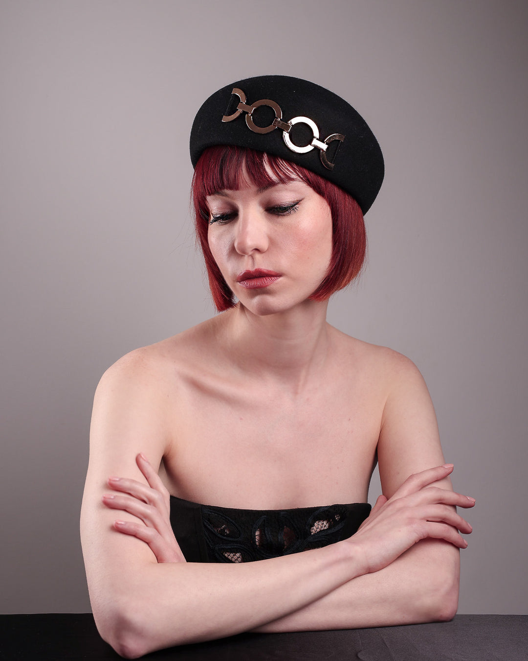 The model showcases the front of the Liora black wool felt beret, which features a geometric gold-tone chain embellishment that arches across the crown. Designed by Merve Bayindir.