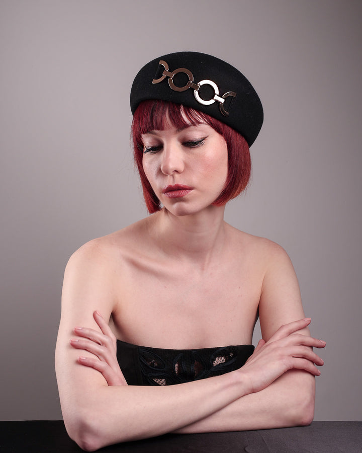 The model showcases the front of the Liora black wool felt beret, which features a geometric gold-tone chain embellishment that arches across the crown. Designed by Merve Bayindir.
