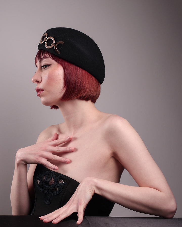 The model showcases the left side from the below angle of the Liora black wool felt beret, which features a geometric gold-tone chain embellishment that arches across the crown. Designed by Merve Bayindir.