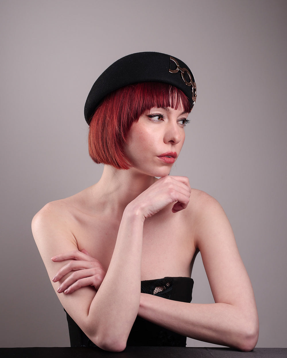 The model showcases the right front of the Liora black wool felt beret, which features a geometric gold-tone chain embellishment that arches across the crown. Designed by Merve Bayindir.
