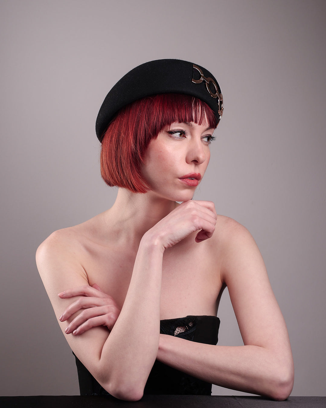 The model showcases the right front of the Liora black wool felt beret, which features a geometric gold-tone chain embellishment that arches across the crown. Designed by Merve Bayindir.