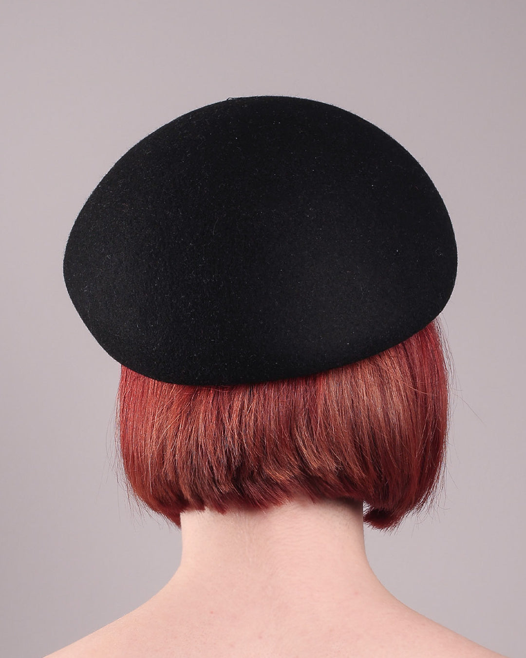 The model showcases the back of the Liora black wool felt beret, which features a geometric gold-tone chain embellishment that arches across the crown. Designed by Merve Bayindir.
