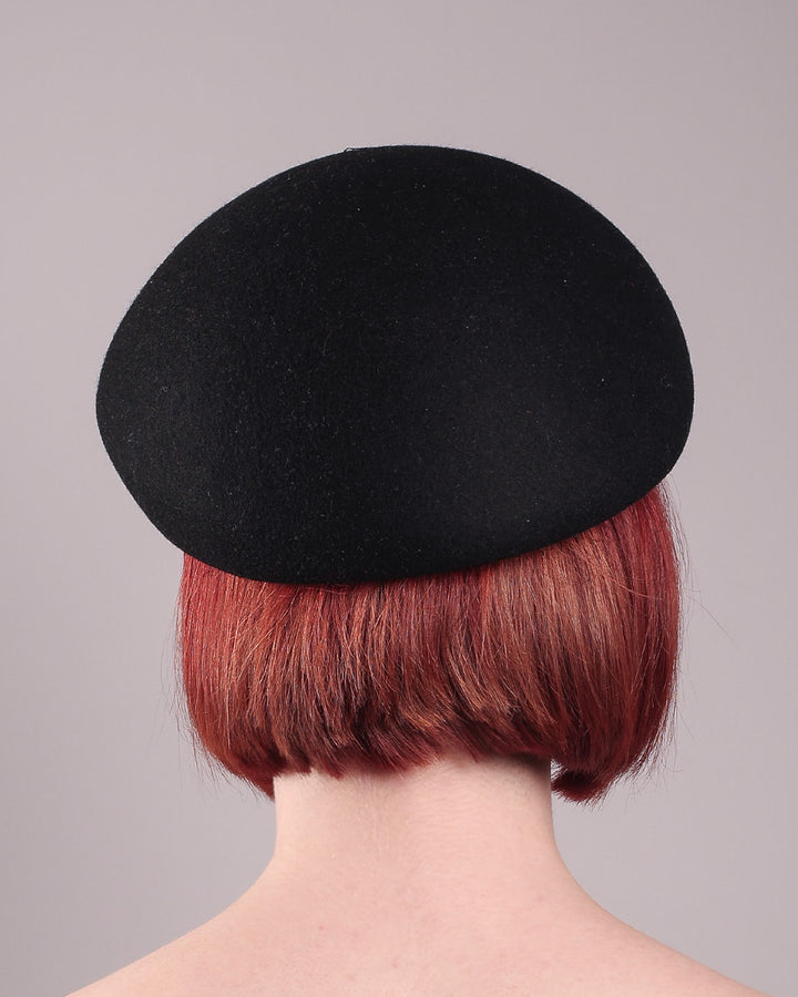 The model showcases the back of the Liora black wool felt beret, which features a geometric gold-tone chain embellishment that arches across the crown. Designed by Merve Bayindir.