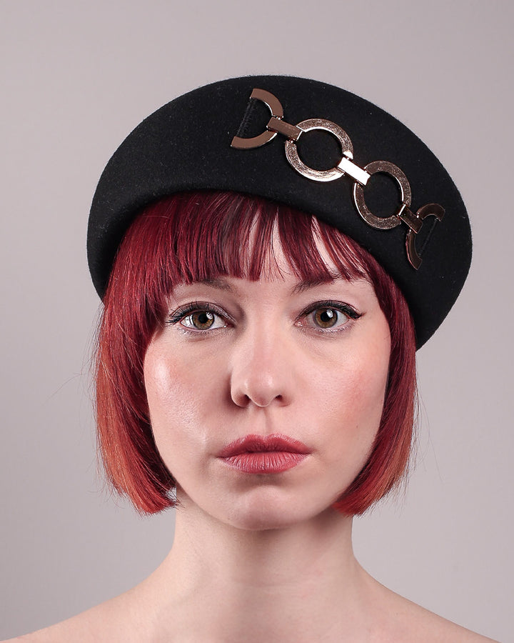 The model showcases the front close up of the Liora black wool felt beret, which features a geometric gold-tone chain embellishment that arches across the crown. Designed by Merve Bayindir.