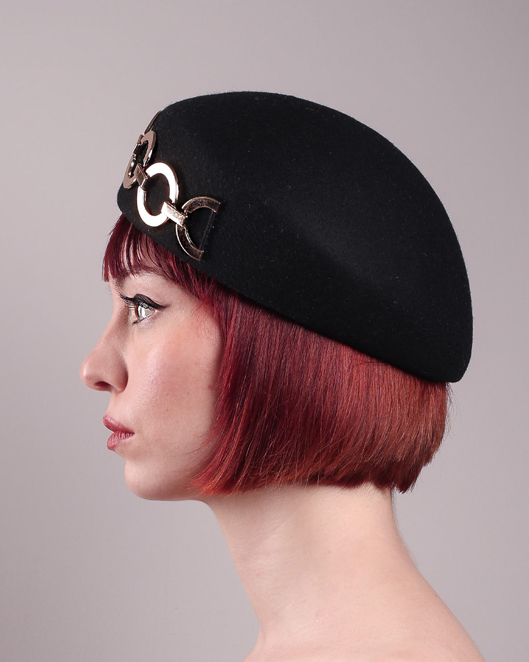 The model showcases the left side close up of the Liora black wool felt beret, which features a geometric gold-tone chain embellishment that arches across the crown. Designed by Merve Bayindir.