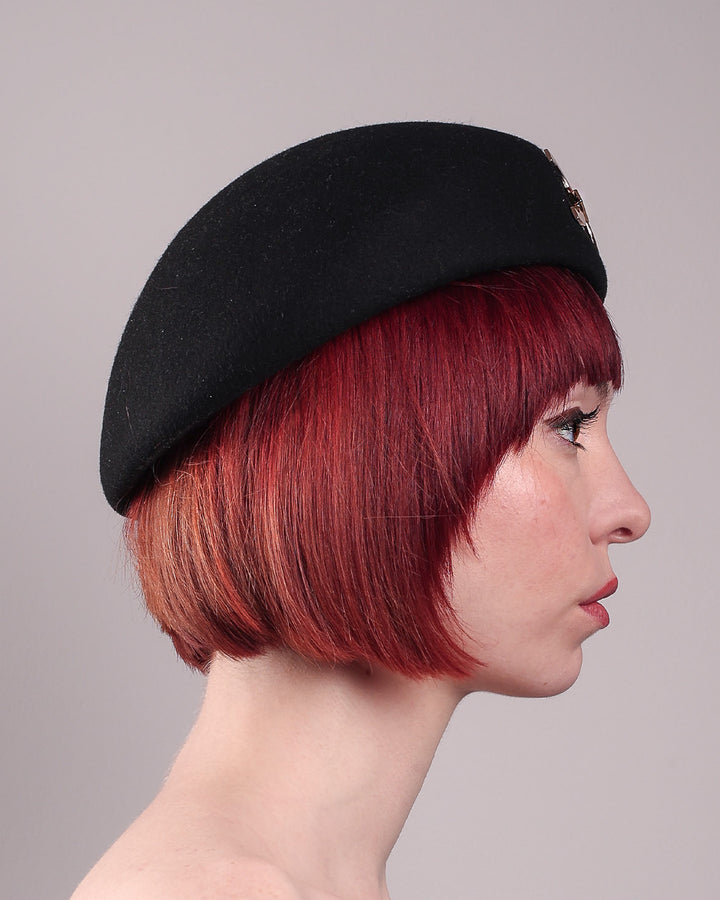 The model showcases the right side close up of the Liora black wool felt beret, which features a geometric gold-tone chain embellishment that arches across the crown. Designed by Merve Bayindir.