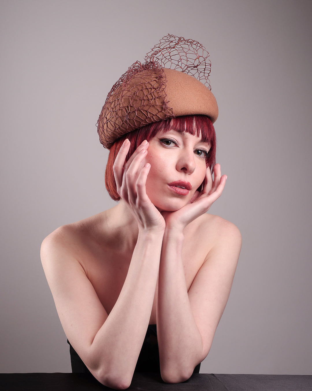 The model is showcasing the right front below angle of the  Lira Pillbox Beret Hat, which is a modern take on classic winter headwear. Crafted from warm brown wool felt, the softly rounded dome shape is elevated by an intricate mesh overlay on the right side, with a fan-like stand on the left side. Designed by Merve Bayindir. 